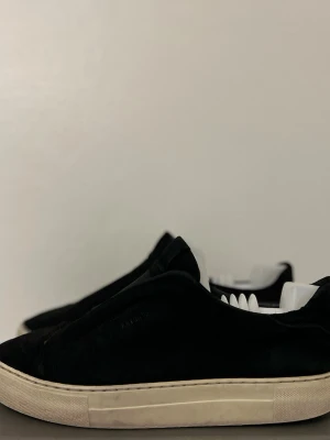 Svarta mockasneakers från Axel Arigato - Säljer ett par svarta sneakers från Axel Arigato i mjuk mocka med vit, platt gummisula. Minimalistisk design utan synlig snörning och diskret logga på sidan. Perfekta för dig som gillar stilrena och cleana skor. Tvättar innan frakt 