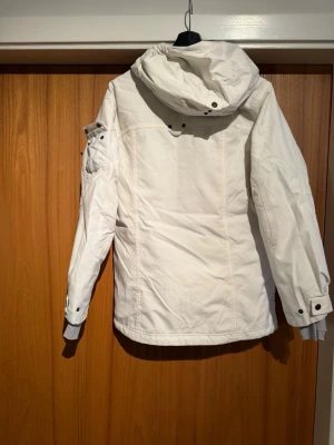 Peak performance Vit parkasjacka  - Säljer en vit parkasjacka från Didriksons med huva och snygga detaljer i läderimitation. Jackan har flera fickor med både knappar och dragkedjor, samt justerbara ärmslut. Perfekt för kalla dagar och riktigt skön att bära.