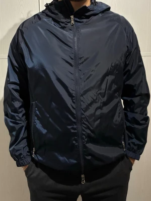 Mörkblå vindjacka J.Lindeberg - Snyggt mörkblå vindjacka från J.Lindeberg med huva och hel dragkedja framtill. Jackan har två sidofickor med dragkedja och elastiska muddar vid ärmslut. Tillverkad i lätt polyester som passar perfekt när det blåser ute. Oanvänd.