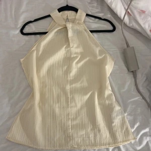 Krämfärgad halterneck topp från SHEIN - Säljer en krämfärgad halterneck topp från SHEIN i storlek S. Toppen har en lätt glansig yta med subtila vertikala ränder och knäppning med pärlknapp i nacken. Materialet är polyester med lite stretch och modellen är ärmlös med hög hals.