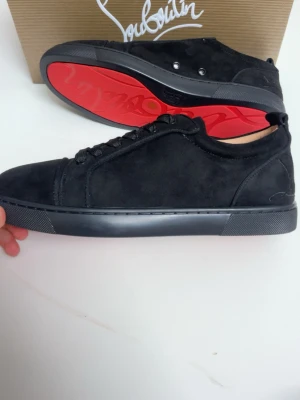 Svarta sneakers från Christian Louboutin - Säljer ett par svarta sneakers från Christian Louboutin med ikonisk röd sula och snygg signatur på hälen. Skorna är i mocka med snörning och har en rund tå. Insidan är beige med guldfärgad logga. Perfekt för dig som gillar lyxiga detaljer och stilren design.