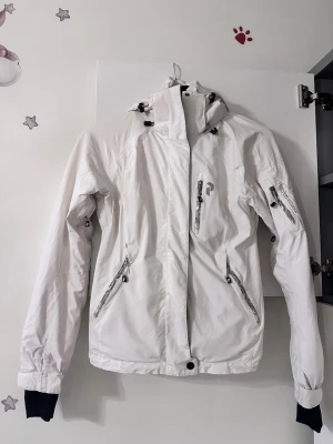 Vit vinterjacka Peak Performance - Säljer en vit vinterjacka från Peak Performance med huva, flera dragkedjeförsedda fickor och snygga detaljer. Jackan har lång ärm, justerbar huva och innerfoder. Perfekt för kalla dagar och har en clean look med diskreta loggor.