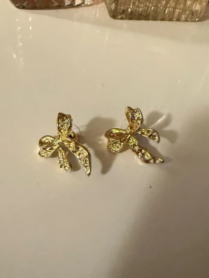 Guld rosettörhängen  - Ett par guldfärgade rosettörhängen från Glitter. Helt nya och oanvända.