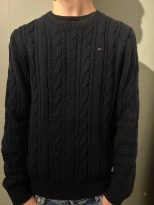 Mörkblå stickad tröja Tommy Hilfiger - Säljer en mörkblå stickad tröja från Tommy Hilfiger med klassiskt kabelstickat mönster. Tröjan har rund hals, långa ärmar och liten broderad logga på bröstet. Ribbstickade muddar vid ärmslut och nederkant. Perfekt för kyliga dagar!