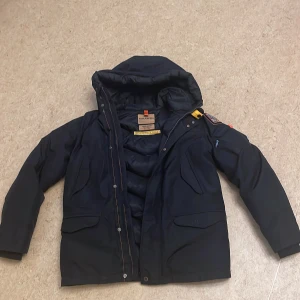 Svart vinterjacka från Parajumpers - Säljer en svart vinterjacka från Parajumpers med huva och dragkedja. Den har använts 3 gånger. Jackan har långa ärmar och flera praktiska fickor. Den är dekorerad med ett broderat märke och knappar för extra stil. Helt äkta köpt från NK Barn avdelning i storleken Young Medium. Passar män med storlek S också. Priset kan diskuteras.