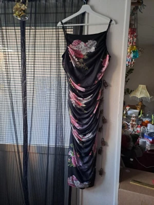 Svart långklänning med blommor - Säljer en svart långklänning med justerbara smala axelband och snygg drapering längs sidorna. Klänningen har ett blommigt mönster i rosa, vitt och grönt som ger en romantisk vibe. Materialet känns lätt och mjukt mot huden, perfekt för dig som gillar att sticka ut.