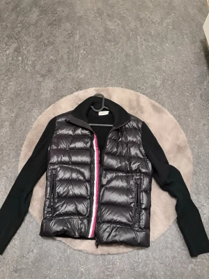 Svart Moncler pufferjacka med stickade ärmar - Säljer en svart pufferjacka från Moncler med stickade långärmade partier och glansig framsida. Jackan har dragkedja med vit och röd detalj, två fickor med dragkedja och Moncler-logga på ärmen. Perfekt för dig som vill ha en snygg och varm jacka med cool mix av material.