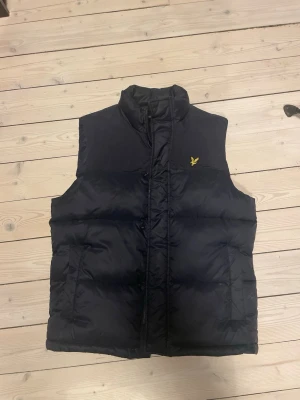 Svart dunväst från Lyle & Scott - Säljer en svart dunväst från Lyle & Scott med gul logga på bröstet. Västen har hög krage, dragkedja och två sidofickor. Perfekt att slänga över en hoodie när det är kyligt ute. Klassisk och stilren design som funkar till många olika outfits.
