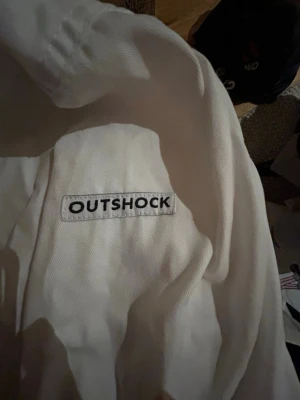Vit kampsportsdräkt från Outshock 170cm - Säljer en vit kampsportsdräkt från Outshock, perfekt för judo eller karate. Setet innehåller jacka, byxor med resår i midjan och ett vitt bälte. Tillverkad i slitstark bomull och passar dig som är runt 165-170 cm lång.