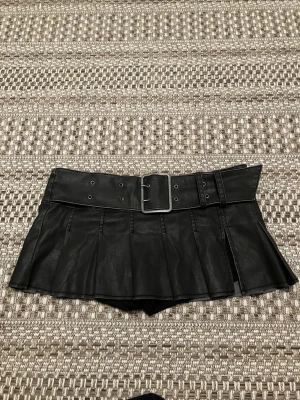 Low waist Minikjol i fejkskinn med bälte - Säljer lowrise mörkgrå mini kjol i fejkskinn med inbyggda shorts, bredt bälte och silverfärgat spänne.  Plisserad/veckad kjoldesign och dragkedja i sidan. Snygg och edgy look som sticker ut. Perfekt till nyår!🌸💕 Använd bara 1 gång därför säljer.