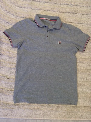 Grå Moncler pikétröja med röd detalj - Säljer en grå pikétröja från Moncler med perfekta ärmar och klassisk krage. Tröjan har tunna röda och vita ränder på krage och ärmslut samt Moncler-logga på bröstet. Materialet är mjuk bomull som känns skönt mot huden. Perfekt för en chill och stilren look. 