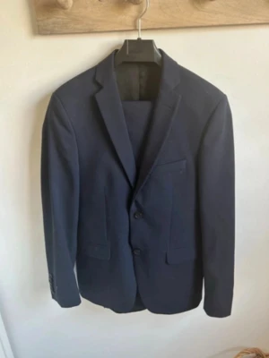 Mörkblå kavaj med klassisk passform - Snygg mörkblå kavaj med två knappar framtill och klassiska fickor. Kavajen har en stilren design med smala slag och är tillverkad i ett slätt material som ger en elegant look. Perfekt för dig som vill ha en enkel och tidlös stil.