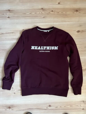 Mörk vinröd sweatshirt från Björn Borg - strl.M - Säljer en mörk vinröd sweatshirt från Björn Borg med trycket 'HEALTHISM' på bröstet. Tröjan har rund hals, långa ärmar och är gjord i mjuk bomull. Perfekt för chill dagar och snygg att matcha med jeans eller joggers. Använd en gång.