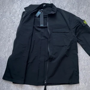 Svart Stone Island overshirt - Svart overshirt från Stone Island med klassisk patch på ärmen. Jackan har dragkedja framtill, bröstficka med lock och stilren krage. Tillverkad i lätt bomullsmaterial, perfekt för lager-på-lager. Snygg och enkel att matcha till streetwear eller casual outfits. (ej äkta)