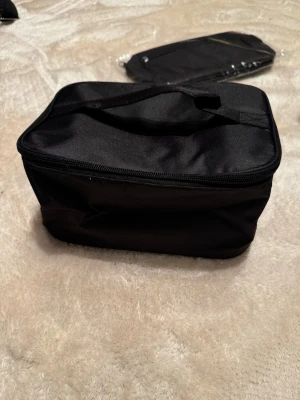 Svart necessär med dragkedja - Praktisk svart necessär med rektangulär form och handtag på ovansidan. Väskan är tillverkad i syntetmaterial och har ett stort huvudfack samt en meshficka med dragkedja på insidan av locket. Perfekt för att organisera dina småsaker eller smink. Helt ny.