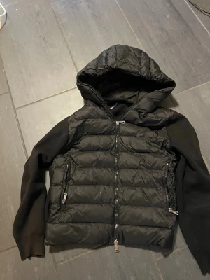 Svart cardigan från Moncler - Svart cardigan från Moncler med stickade ärmar och rygg. Jackan har huva, två sidofickor med dragkedja och quiltad framsida. Materialmix av polyester och stickad ull ger en schysst kontrast. Perfekt för dig som gillar streetstyle och vill ha något som sticker ut.