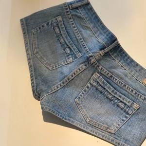Low waist jeans shorts - Snygga blå low waist jeansshorts med dekorativa banddetaljer på både fram- och bakfickor. Klassisk femficksmodell med bälteshällor och knappstängning. Perfekta för varma dagar och enkel att matcha med olika toppar.
