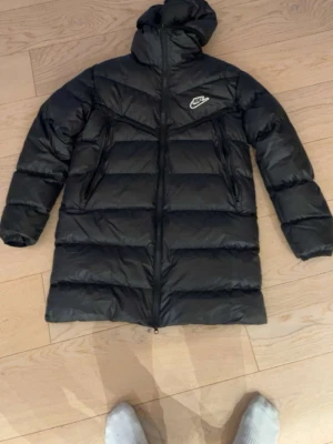 Svart lång dunjacka från Nike - Säljer en svart lång dunjacka från Nike med huva och dragkedja hela vägen. Jackan har en quiltad design och två sidofickor med dragkedja. Perfekt för kalla dagar och riktigt skön att bära. Klassisk Nike-logga på bröstet.