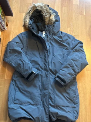 Grå parkajacka med fuskpäls från McKinley - Parkajacka från McKinley i grått med stor huva och fuskpäls. Jackan har långa ärmar, stora fickor framtill och justerbara muddar med kardborre. Perfekt för kalla dagar och har en stilren look som funkar till många outfits.