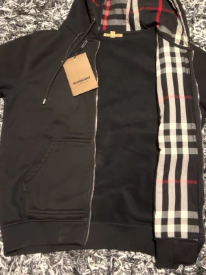 Svart Burberry hoodie - Svart hoodie från Burberry i storlek M, tillverkad i 100% bomull. Huva med klassiskt Burberry- rutigt mönster i svart, vit, grå och röd. Dragkedja framtill, störning vid huvan och två fickor. Perfekt för dig som gillar stilrena streetwear- vibbar.