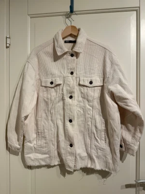 Vit manchesterjacka från Zara - Säljer en oversized vit manchesterjacka från Zara med svarta knappar och två bröstfickor. Jackan har en rå kant nertill och breda ribbor i tyget.