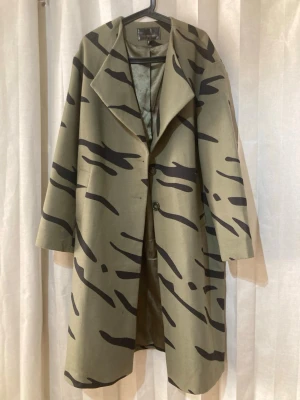 Camouflageinspirerad kappa  - Snygg kappa i olivgrönt med svart camouflage-mönster. Loose passform med bred krage, helfodrad och två knappar framtill. En statement-jacka för dig som vill sticka ut och bygga en cool, avslappnad look. Perfekt till både vardag och streetstyle. Helt ny köptes för 2590 kr 