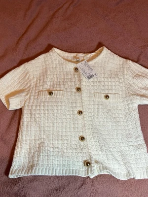 Vit stickad kofta med kort ärm och guld detaljer  - Vit stickad kofta med guld detaljer och Ny med prislapp 