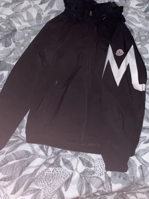 Svart vindjacka från Moncler - Säljer en svart vindjacka från Moncler med stor vit M-detalj på ärmen och klassisk Moncler-logga på bröstet. Jackan har huva, dragkedja framtill och är tillverkad i ett lätt, syntetiskt material. Perfekt för dig som vill ha en clean och sportig look.