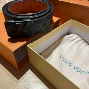 Louis Vuitton bälte - Säljer en stilren svart väst från Louis Vuitton med diskret rutigt mönster. Västen har en klassisk form och är tillverkad i exklusivt skinnmaterial. Perfekt för dig som vill ha en lyxig och tidlös look med ikonisk LV-detalj framtill. Bältet är äkta!