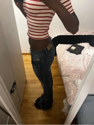 Blå Diesel jeans med slitningar - Säljer ett par blå jeans från Diesel med snygga slitningar och tvättade detaljer. Modellen har låg midja och bootcut med klassiska fickor både fram och bak. Jeansen har vit kontrastsöm och en Diesel-patch bak i midjan. Perfekt för dig som gillar en cool och avslappnad stil.