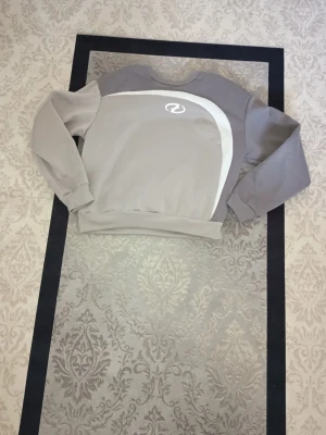 Grå sweatshirt - Säljer en stilren grå sweatshirt med vit logga och vit böjd detalj på bröstet. Tröjan har långärmade muddar och en klassisk rund hals. Perfekt för chill dagar eller när du vill ha en enkel look.