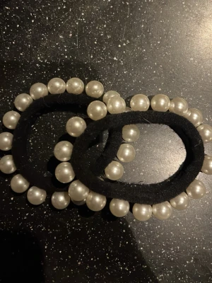 Svarta hårsnoddarmed pärldetaljer - Säljer två svarta hårsnoddar med snygga pärlor längs kanterna som ger en lyxig och trendig vibe. Perfekt för dig som vill sticka ut och addera lite extra bling till outfiten. Hårsnoddarma har en enkel form och pärlorna är vita och runda, vilket gör den superenkel att matcha.