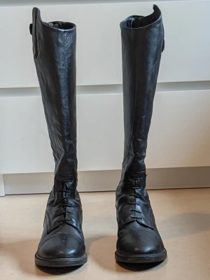 Ridstövlar från CRW i skinn - Säljer ett par höga svarta ridstövlar från CRW i äkta skinn. Klassisk design med snörning framtill och rund tå. Perfekta för dig som vill ha stilrena och funktionella stövlar till ridning. Metallmärke med CRW-logga på sidan. Två små defekter på utsidan av ridstövlarna, visas på bild