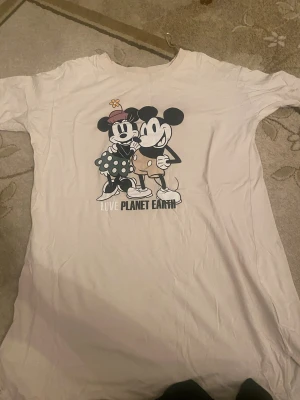 Vit Disney t-shirt med Mickey & Minnie - Säljer en vit t-shirt från C&A i samarbete med Disney, storlek L. Framsidan har ett stort tryck med Mickey och Minnie Mouse och texten 'LOVE PLANET EARTH'. T-shirten är kortärmad och gjord i mjuk bomull, perfekt för dig som gillar retro och tecknade motiv.