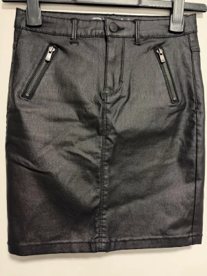 Svart kortkjol med dragkedjor ONLY - Snygg svart kortkjol i skinnimitation med två framfickor med dragkedjor och bakfickor. Kjolen har bälteshällor och knapp framtill.