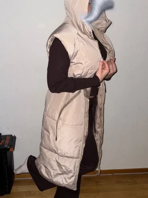 saint tropez Väst HellaSZ Vest - Säljer en beige lång dunväst med huva och dragsko i midjan. Västen har en quiltad design och dragkedja framtill. Perfekt att slänga över en hoodie för extra värme. Fickor på sidorna och snygg, loose passform. Nypris 1299kr 