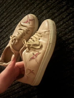 Rosa axel arigato skor med broderade fåglar - Säljer ett par rosa axel arigato sneakers med snörning och rund tå. Skorna har broderade fåglar i rosa och svart på sidorna och framtill, vilket ger dem en unik och cool look. Tillverkade i skinn med platt sula och neutrala färger som passar till många outfits. (Har tvättat dem så de är mycket renare än på bilderna) skriv för fler bild