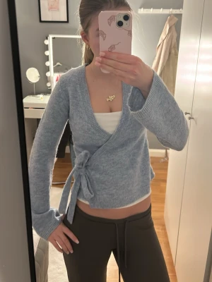 Ljusblå omlottkofta från Gina Tricot - Supermysig ljusblå omlottkofta från Gina Tricot i ullblandning. Koftan har långa ärmar, v-ringning och knyt i sidan för en snygg och avslappnad look. Perfekt att slänga på sig när det är lite kyligt ute.