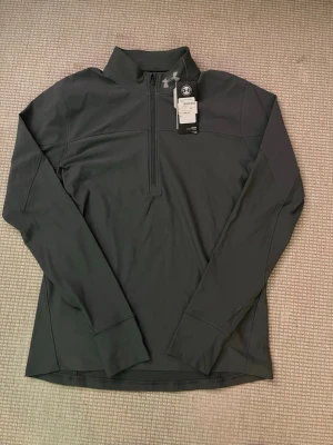Svart träningströja från Under Armour - Snygg svart långärmad träningströja från Under Armour med halv dragkedja framtill och hög krage. Tröjan är gjord i ett mjukt syntetmaterial som andas och har en diskret logga på bröstet. Perfekt för gymmet eller löprundan. Originella pris är 499kr kommer sälja den för 300kr
