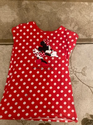 Röd prickig topp med Minnie Mouse - Säljer en röd topp från Disney i storlek L med vita prickar och ett stort tryck av Minnie Mouse på framsidan. Toppen har rund hals och är ärmlös, tillverkad i mjuk bomull. Perfekt för dig som gillar färgglada och lekfulla plagg!