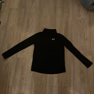 Svart långärmad träningströja Under Armour - Svart långärmad träningströja från Under Armour med halv dragkedja och logga på bröstet. Modellen är loose fit och passar perfekt för sport eller gym. Materialet är syntet och tröjan har en stilren design som funkar till alla träningspass.