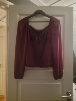 Vinröd omlottblus  - Säljer en vinröd blus från COS med omlottdetalj och knyt framtill. Blusen har långärmade puffiga ärmar och en mjuk, slät yta. Modellen är croppad och har en snygg v-ringning med rynkade detaljer vid bysten. Perfekt för dig som gillar stilrena och trendiga plagg.