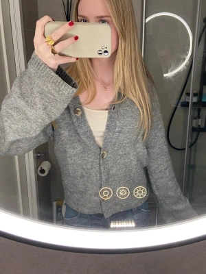 Grå stickad kofta med knappar - Mysig grå stickad kofta med stora knappar framtill. Koftan har en loose passform och långa ärmar, perfekt att slänga på sig över ett linne eller t-shirt. Enkel och stilren design som funkar till jeans eller kjol.