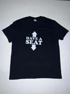 ”Have a seat” t-shirt - En svart t-shirt med ett enkelt och humoristiskt tryck. Texten “Have a seat” kompletteras av pilar uppåt och nedåt, vilket ger ett lekfullt budskap. Perfekt som rolig vardagströja eller till festliga sammanhang. Tröjan är i nytt skick och bekväm att bära.
