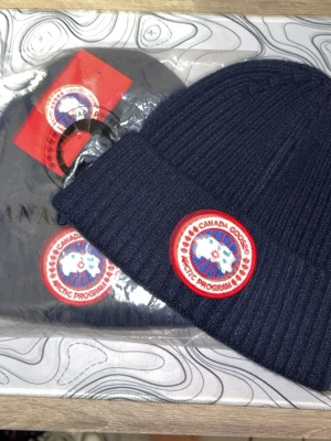 Canada Goose ribbad mössa i olika färger - Snygg ribbstickad mössa från Canada Goose i marinblått med klassisk rund logopatch framtill. Mössan är mjuk och har en bred uppvikt kant för extra stilpoäng. Perfekt för kalla dagar och enkel att matcha med jacka eller hoodie. I den nysvenska grisch stilen!