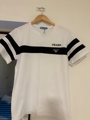 Vit och svart t-shirt från Prada - Snygg vit t-shirt från Prada med svarta ränder över bröstet och på ärmarna. T-shirten har broderad Prada-logga och metallmärke på framsidan. Rund hals och korta ärmar, tillverkad i mjuk bomull. Perfekt för dig som gillar stilrena designer och exklusiva märken.