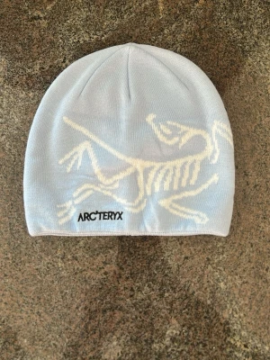 Ljusblå mössa från Arc'teryx - Snygg ljusblå mössa från Arc'teryx med vit dinosaurie-tryck på framsidan och logga. Mjuk och stretchig, perfekt för kyliga dagar. Passar dig som gillar stilrena accessoarer med cool detalj.