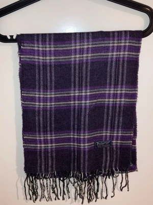 Lila rutig Burberrys halsduk - Säljer en lila rutig halsduk från Burberrys, tillverkad i 100% lammull. Halsduken har klassiskt rutmönster i lila, grått och vitt samt fransar längst ner.