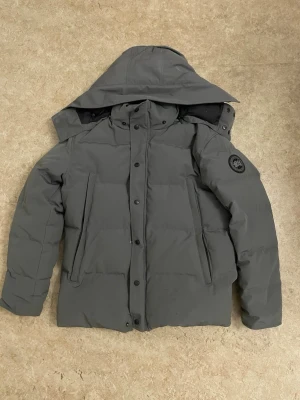 Grå pufferjacka från Canada Goose - Säljer en grå pufferjacka från Canada Goose med huva och svarta detaljer. Jackan har en stilren look med raka linjer, två sidofickor med dragkedja och logotyp på ärmen. Perfekt för kalla dagar och har en skön, vadderad känsla.