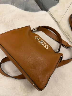 Brun axelväska från Guess - Snygg brun axelväska från Guess i strukturerat skinn med metallfästen och logga framtill. Väskan har både handtag och justerbar axelrem, samt ett smart lås i metall. Perfekt storlek för att få plats med allt du behöver och stilren design.